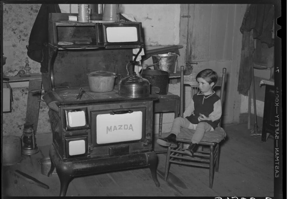 Child sitting beside a cast-iron kitchen cookstove in Vermont in 1939, illustrating how Americans kept warm during winter.