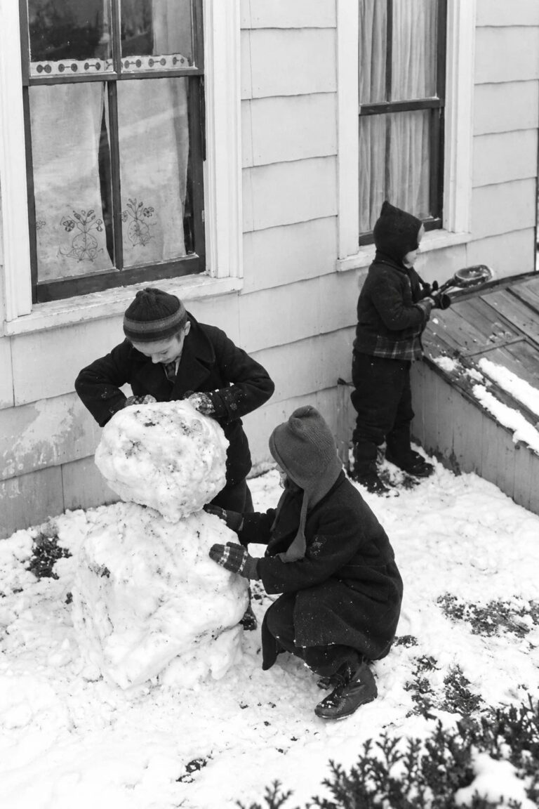Three children wearing warm winter coats, knitted hats, and mittens build a snowman beside an old-fashioned American home.